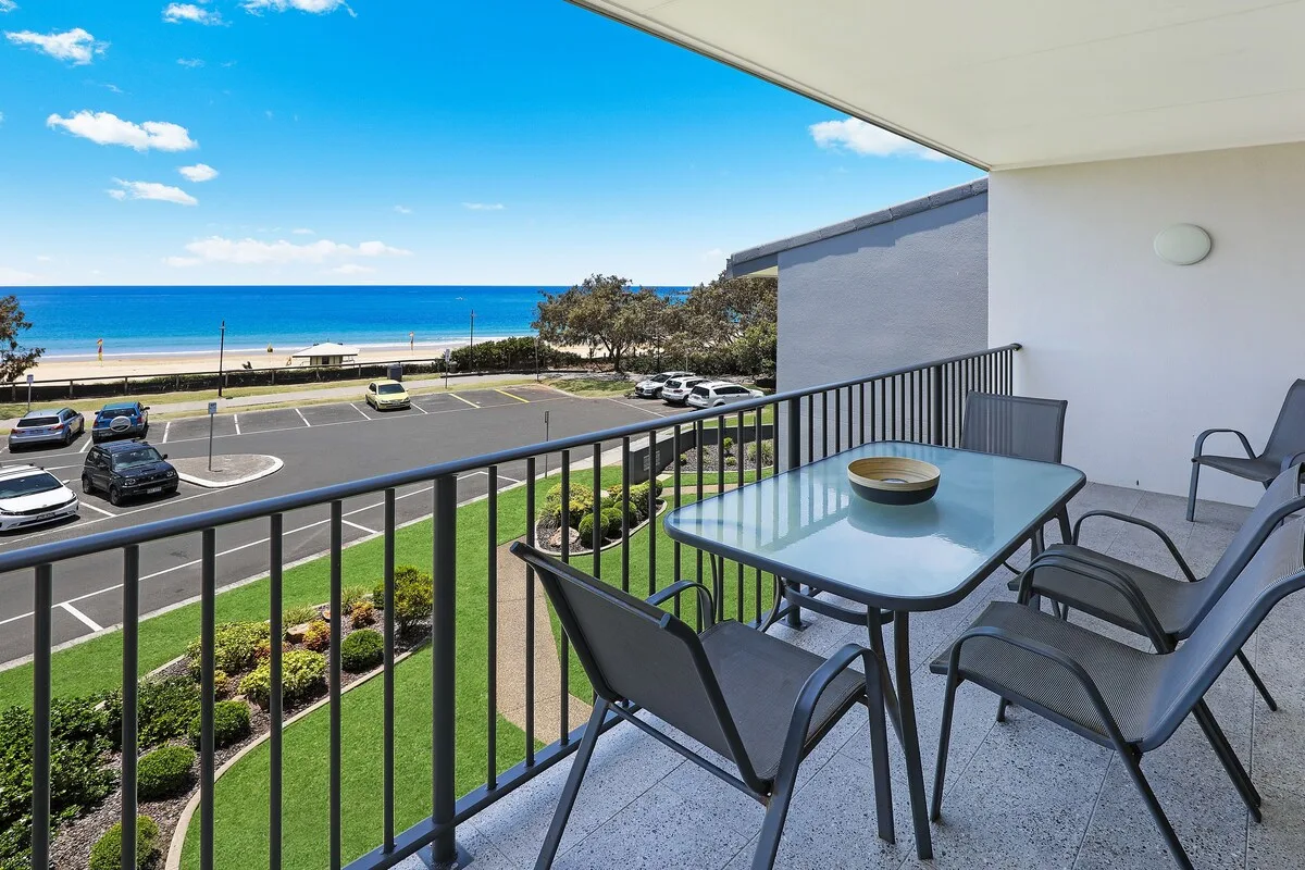A balcony with outdoor seating overlooks a parking area and the ocean.