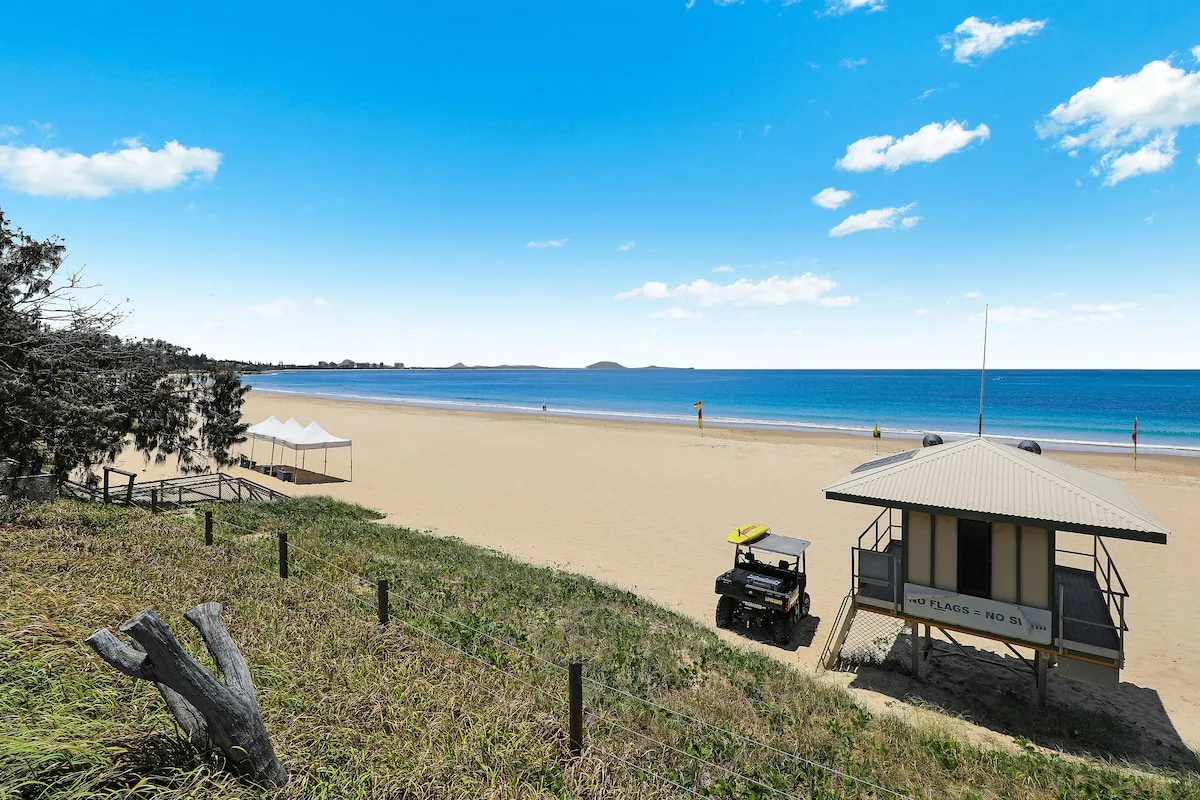 A sandy beach and blue ocean stretch beneath a bright, partly cloudy sky.