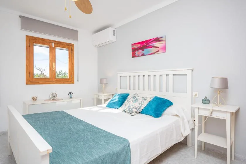 Un dormitorio luminoso con una cama blanca, ropa de cama verde azulado y una ventana con marco de madera.