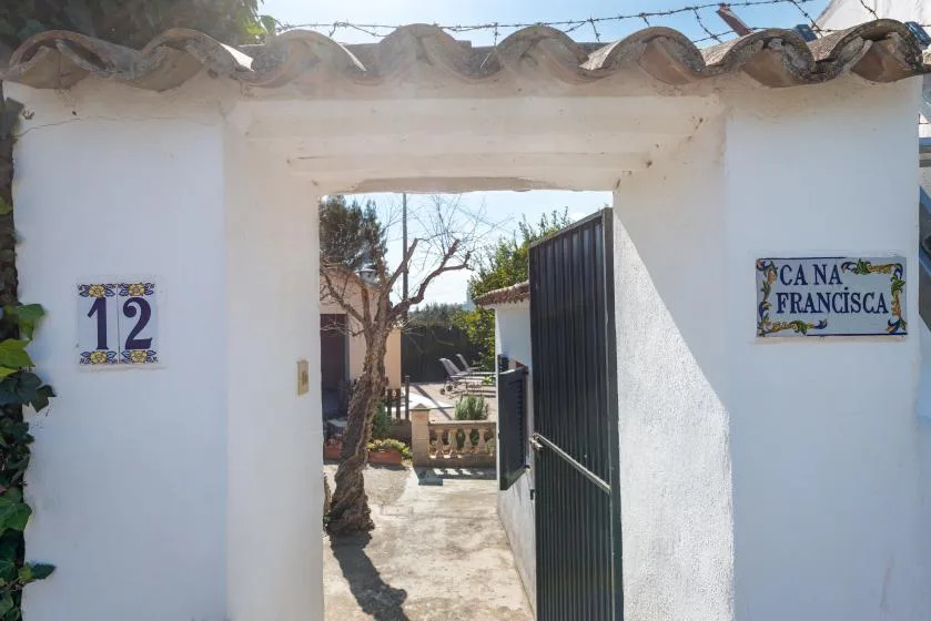 Un muro de entrada blanco tiene una puerta oscura abierta hacia un patio soleado.