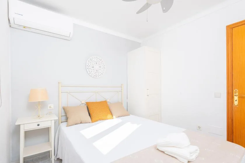 Un dormitorio luminoso con una cama blanca, paredes claras y una puerta de madera.