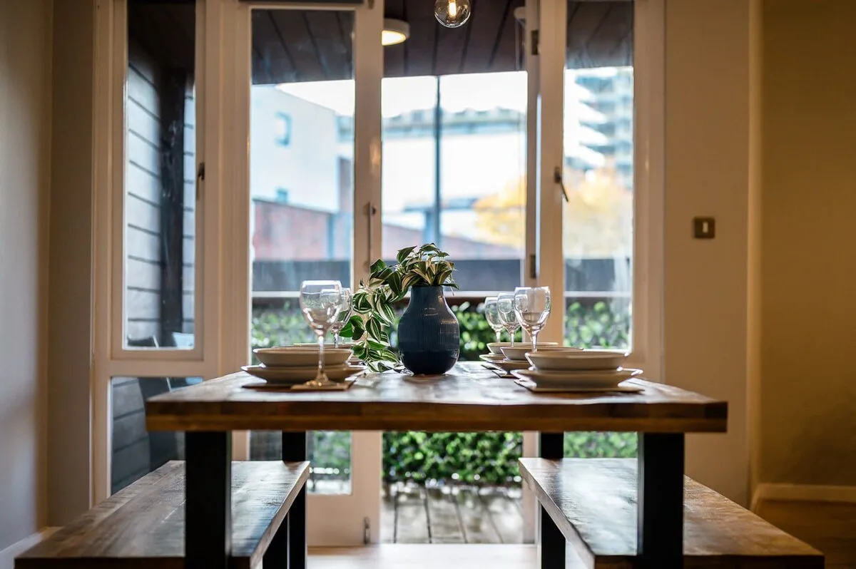 Una mesa de comedor está puesta con platos y cristalería frente a grandes ventanas.