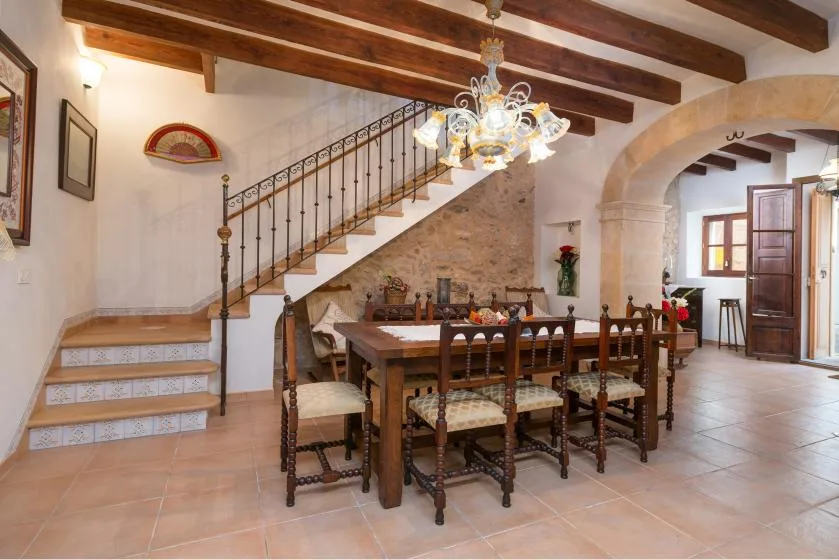 A dining room with a long wooden table beneath exposed ceiling beams and a staircase.