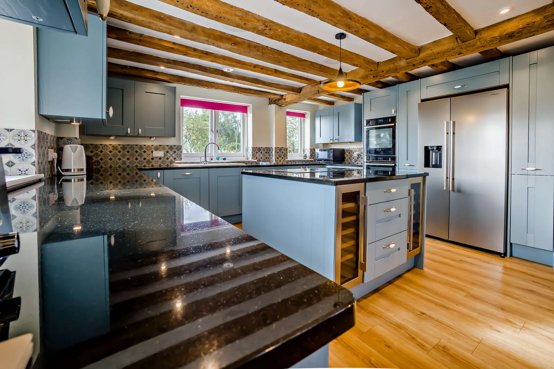A spacious kitchen with blue cabinetry, dark countertops, and exposed wooden ceiling beams.