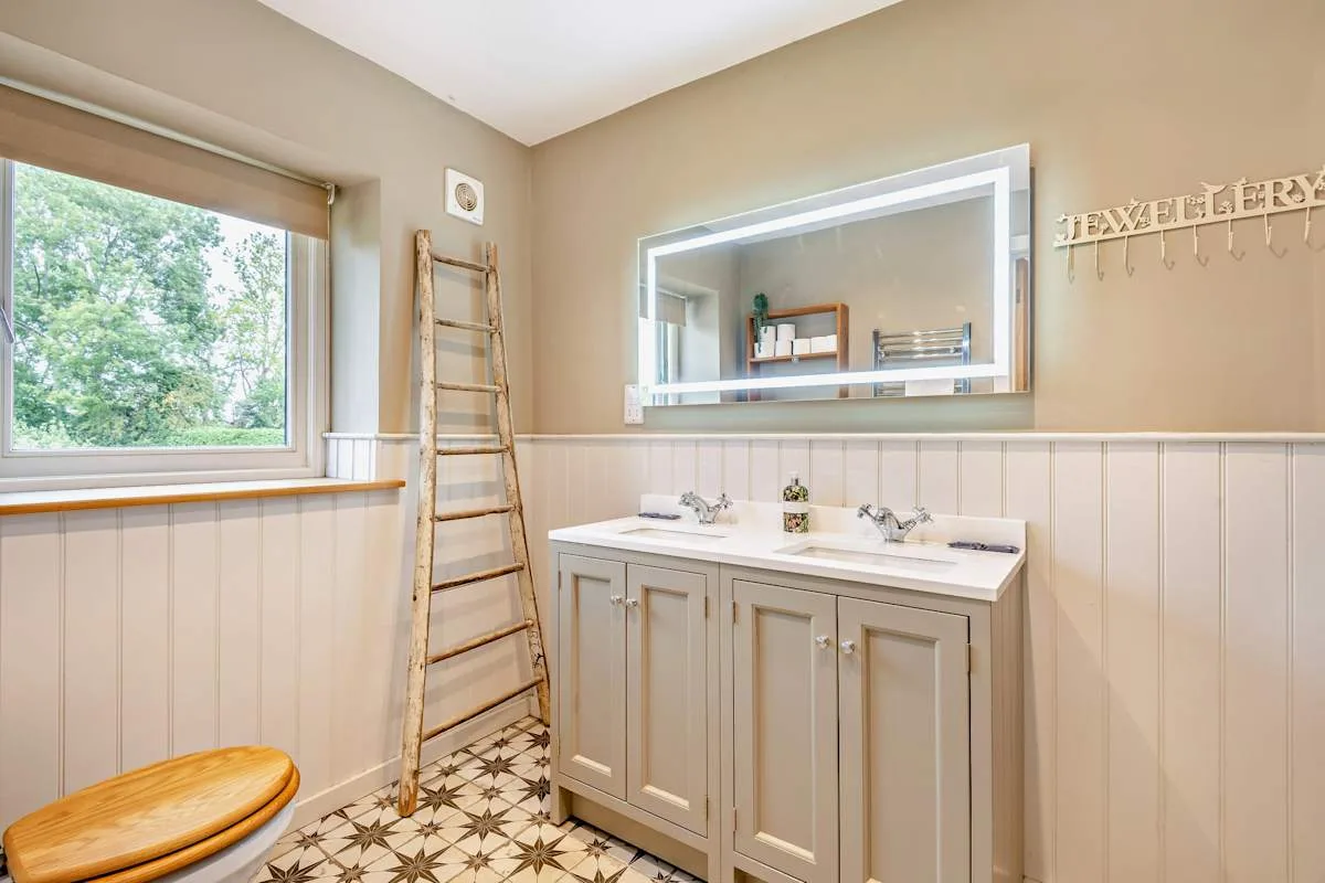 A bathroom with a double vanity, wall mirror, and window overlooking trees.