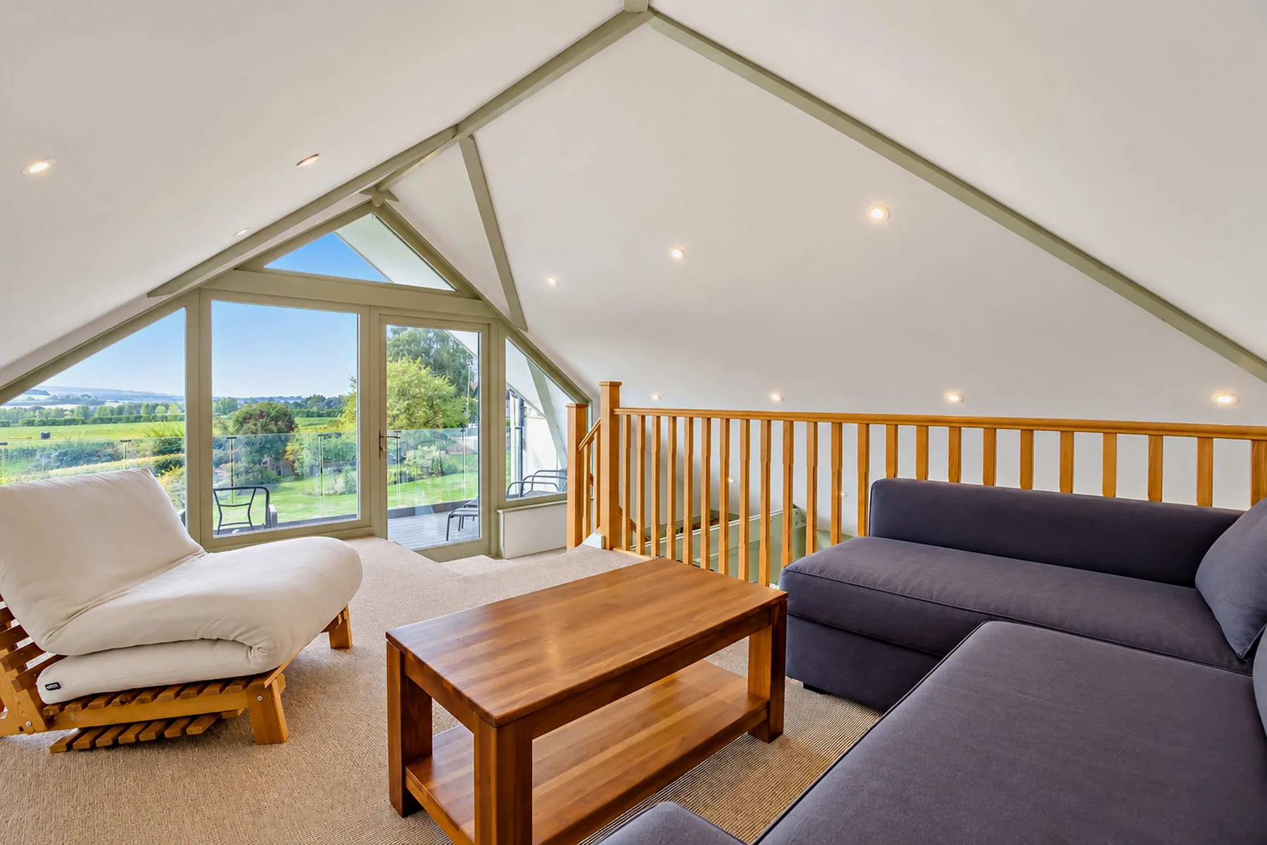 A bright loft sitting area has a sectional sofa, wooden coffee table, and large windows.