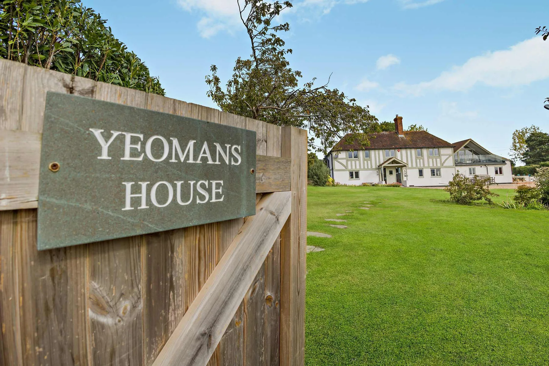 A white country house stands beyond a wooden gate and broad green lawn.