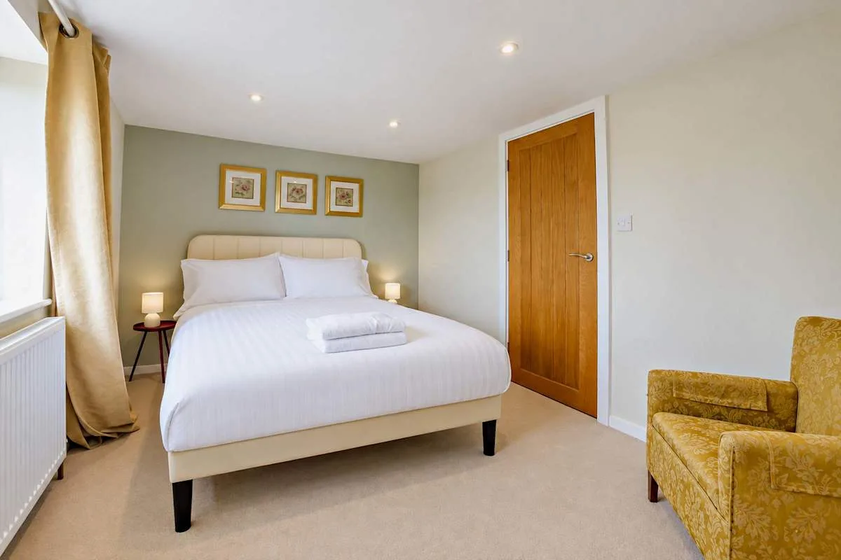 A bedroom with a neatly made bed and a mustard-colored armchair.