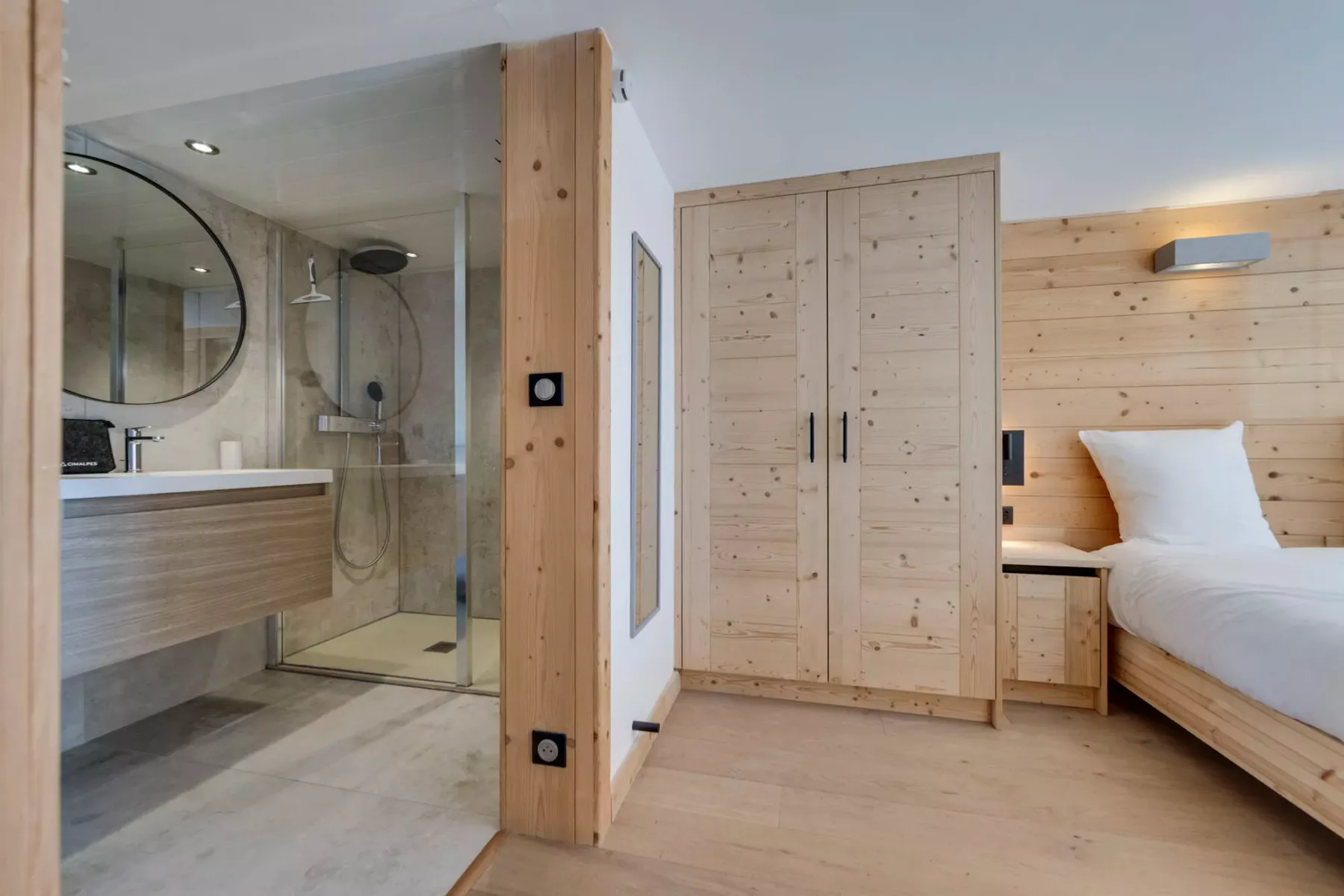 Un dormitorio con muebles de madera clara y una ducha acristalada contigua.