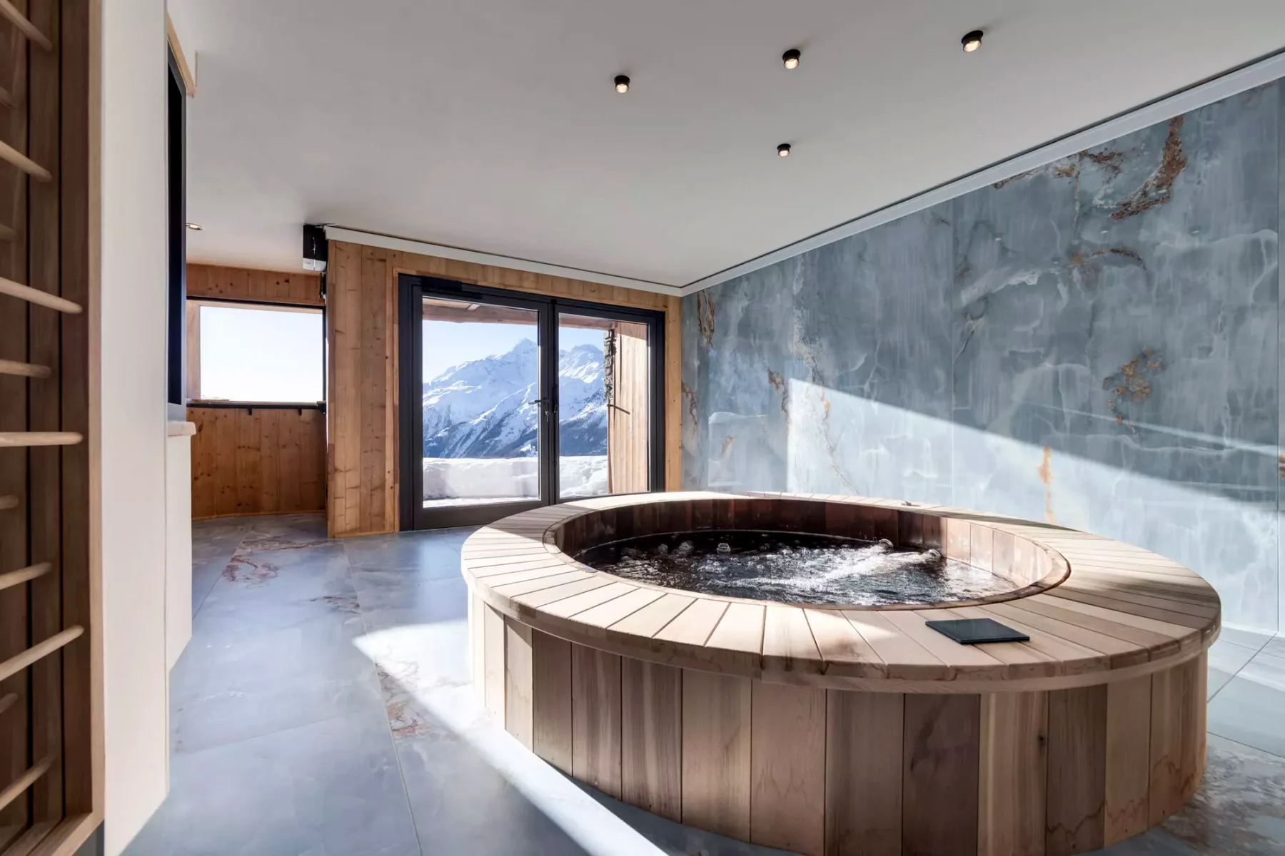 Un spa interior cuenta con una bañera circular junto a grandes ventanales con vistas a montañas nevadas.