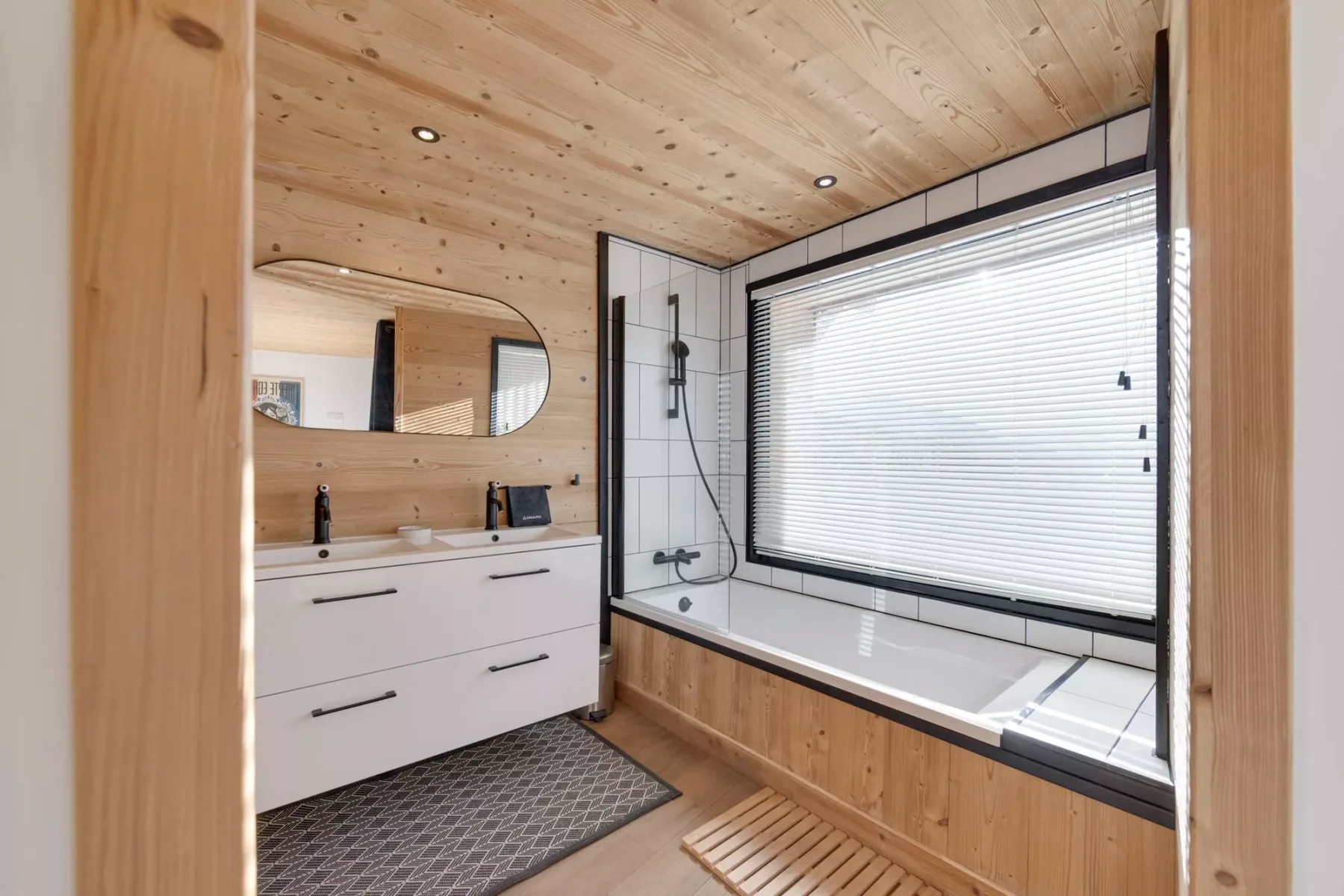 Un baño revestido de madera con lavabo y bañera bajo una ventana grande.