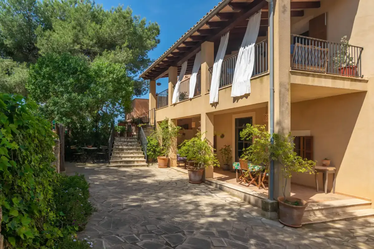 Un patio exterior soleado se extiende junto a un edificio de dos plantas con balcones y plantas en macetas.