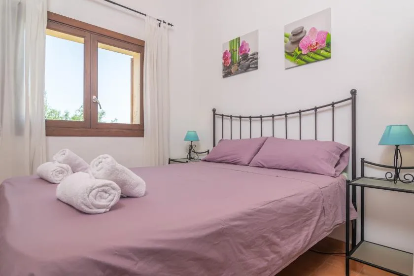 Un dormitorio con una cama cubierta de color malva, mesitas de noche y una ventana.