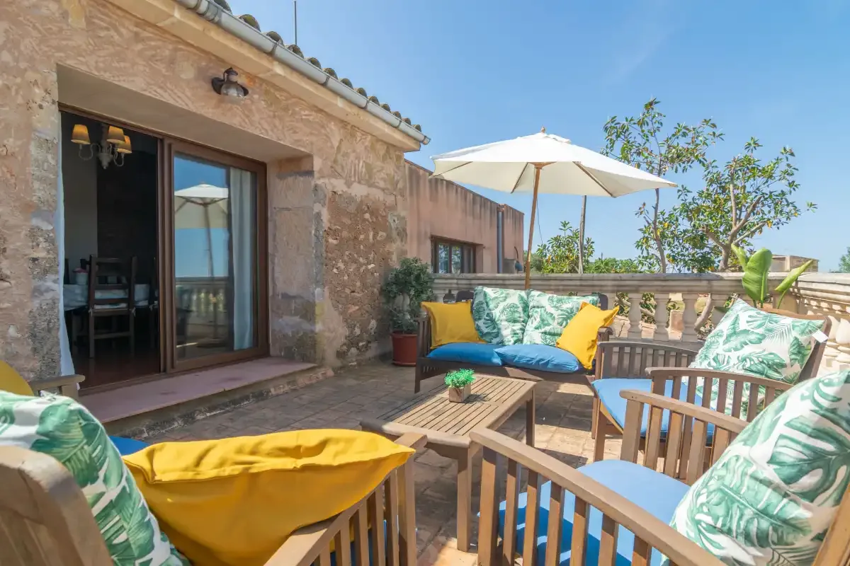 Un patio soleado tiene asientos con cojines, una mesa baja y una sombrilla grande.