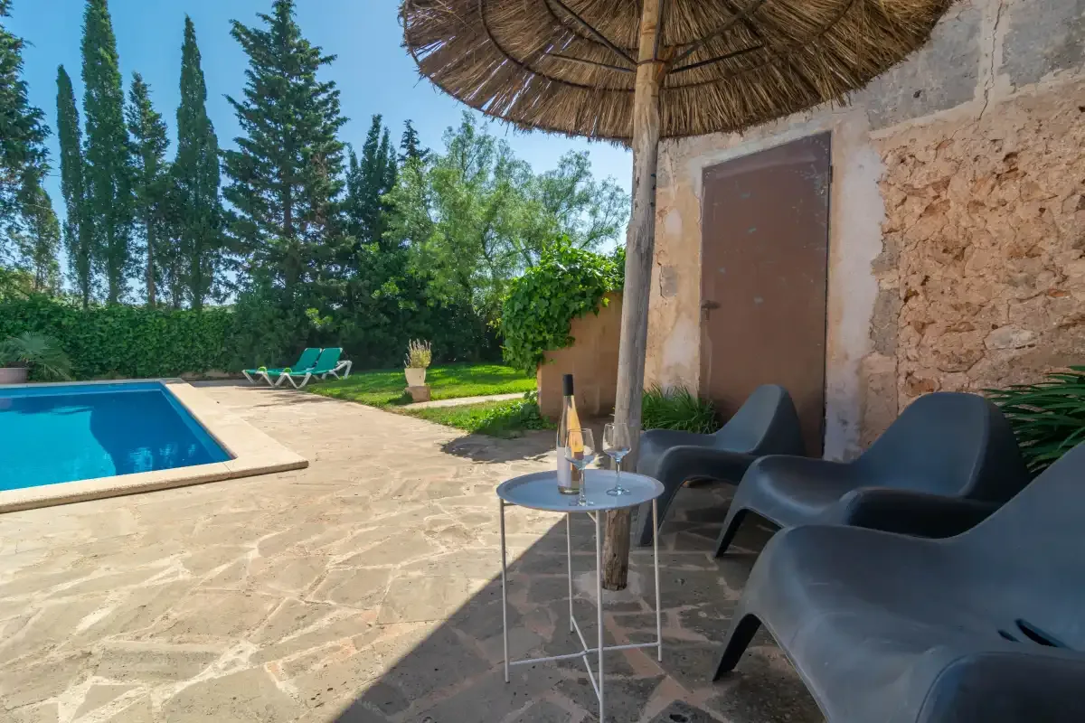 Una piscina junto a un patio de piedra con asientos al aire libre y una sombrilla de paja.