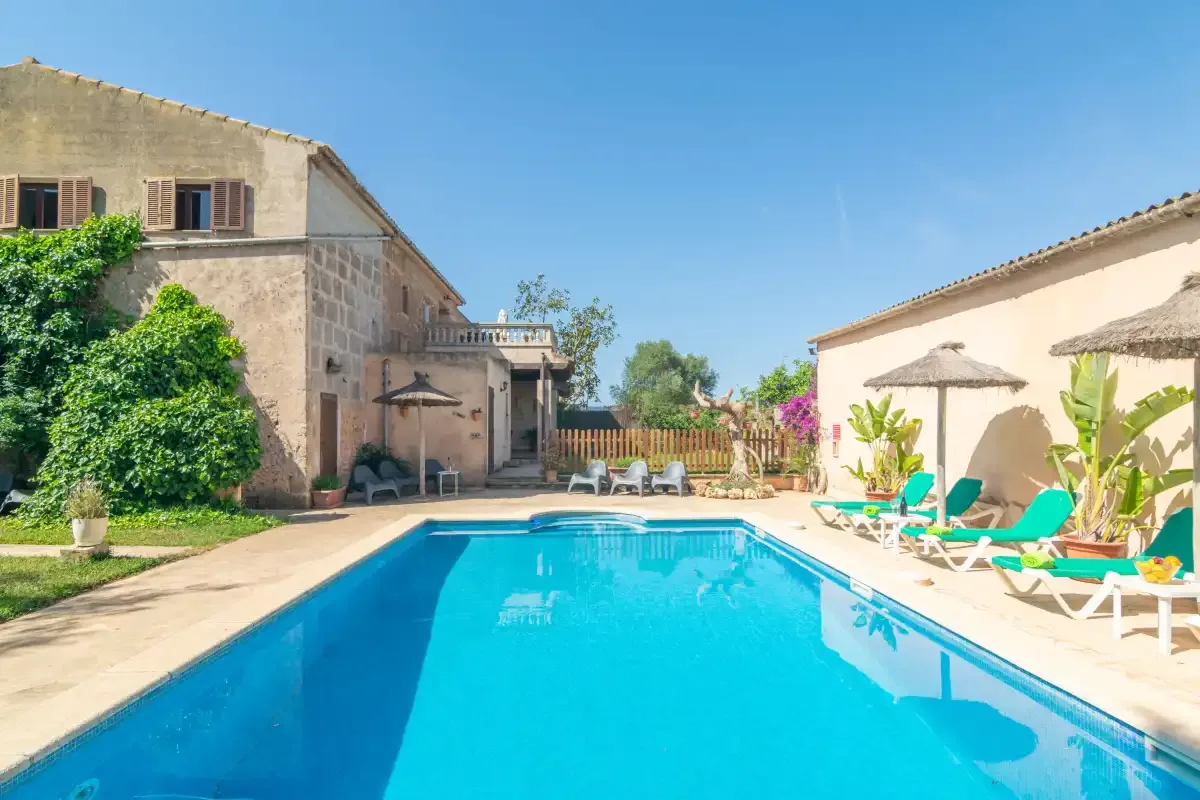 Una piscina exterior se encuentra entre una casa de piedra y un patio con tumbonas.