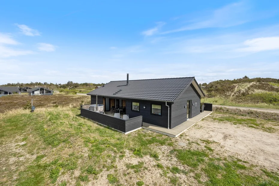 A dark-sided house sits in a grassy, sandy landscape beneath a clear sky.