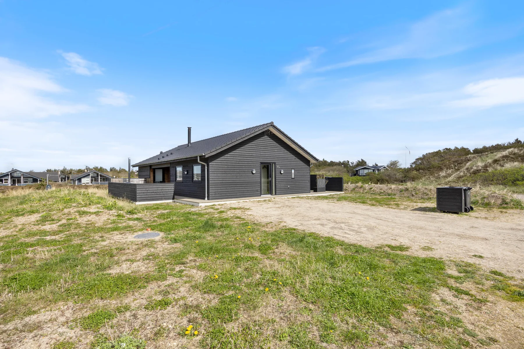 A dark-sided house stands in an open sandy yard beneath a blue sky.