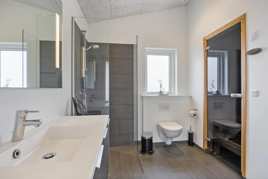 A bright bathroom with a wide sink, wall-mounted toilet, and glass shower screen.