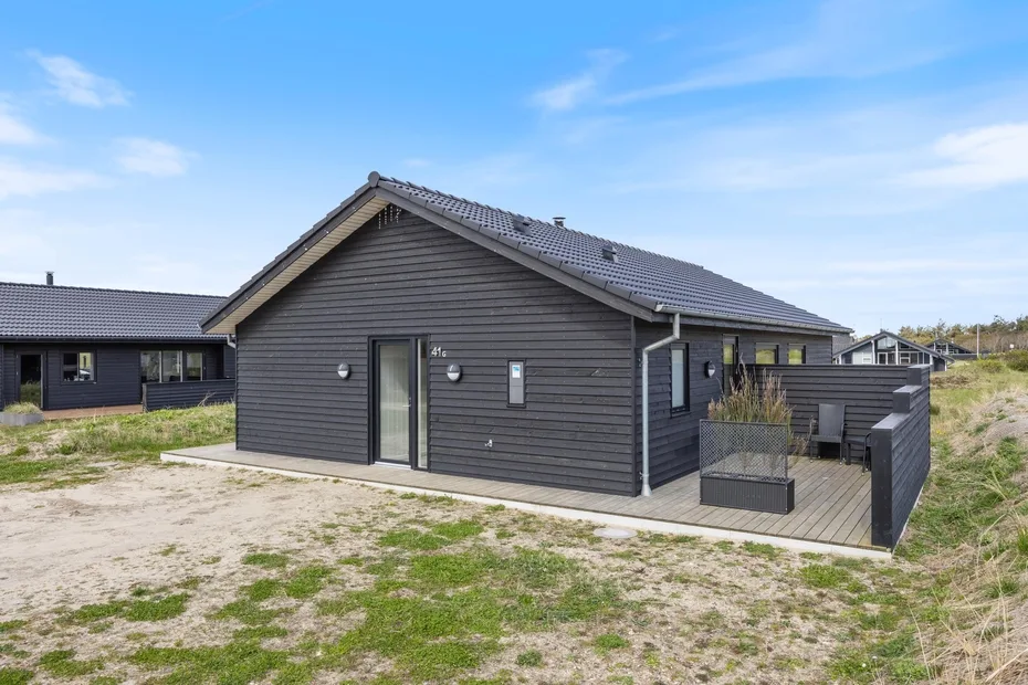 A dark-sided house with a small wooden deck sits in an open sandy landscape.