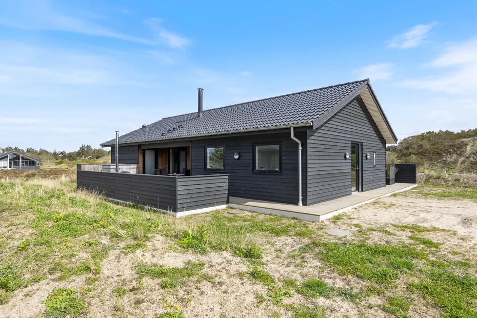 A dark-sided house stands on a grassy, sandy plot beneath a clear sky.