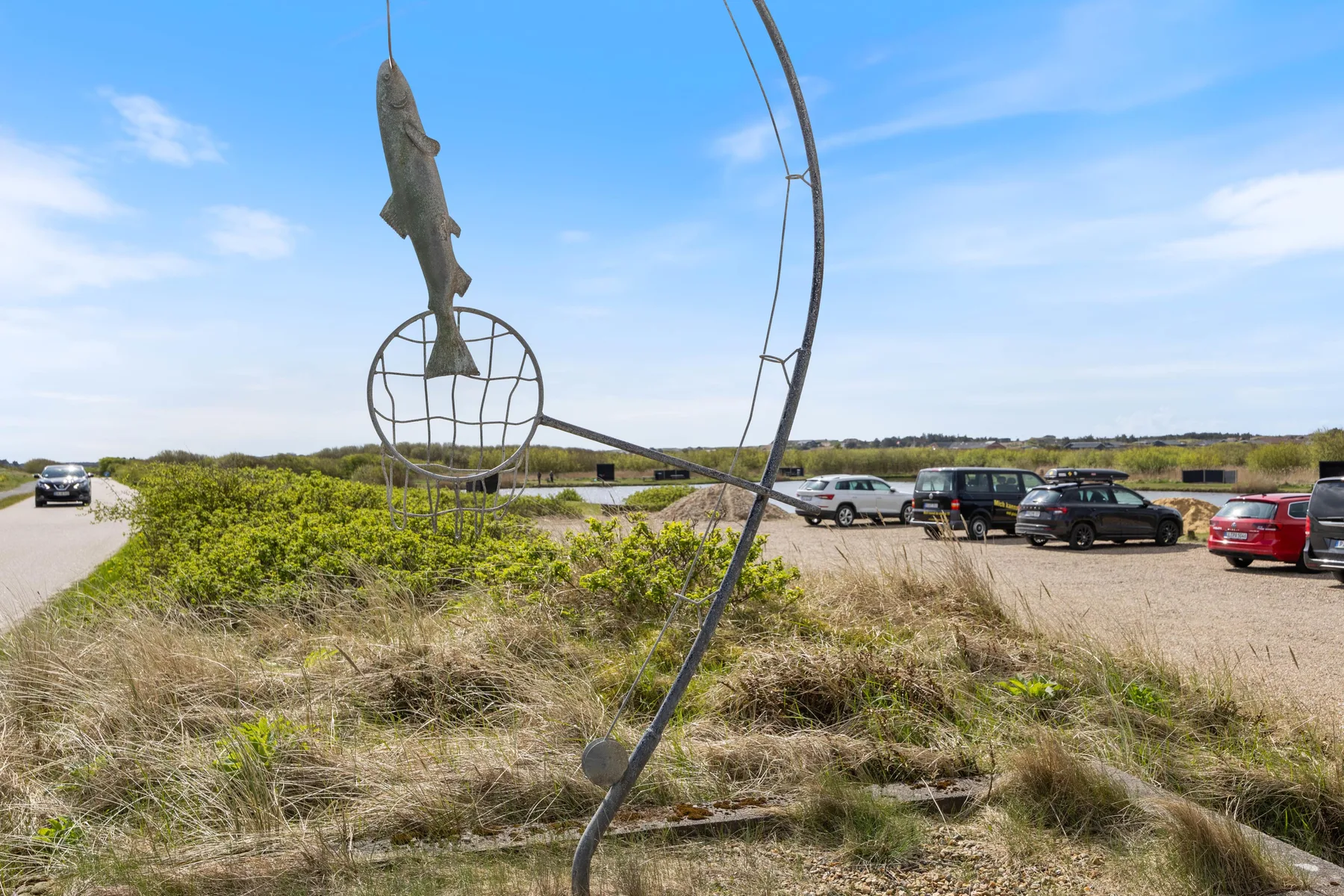 A roadside parking area sits beside grassy dunes and a large fish sculpture.