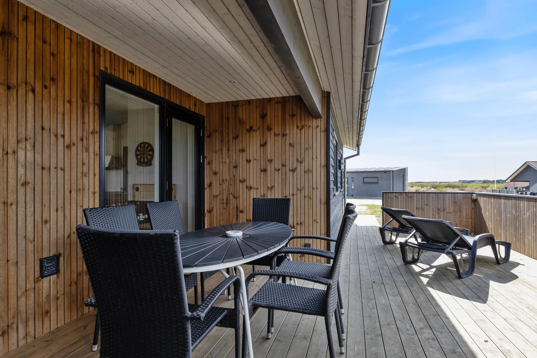 A wooden outdoor deck has a dining table and seating beside a sunny open terrace.