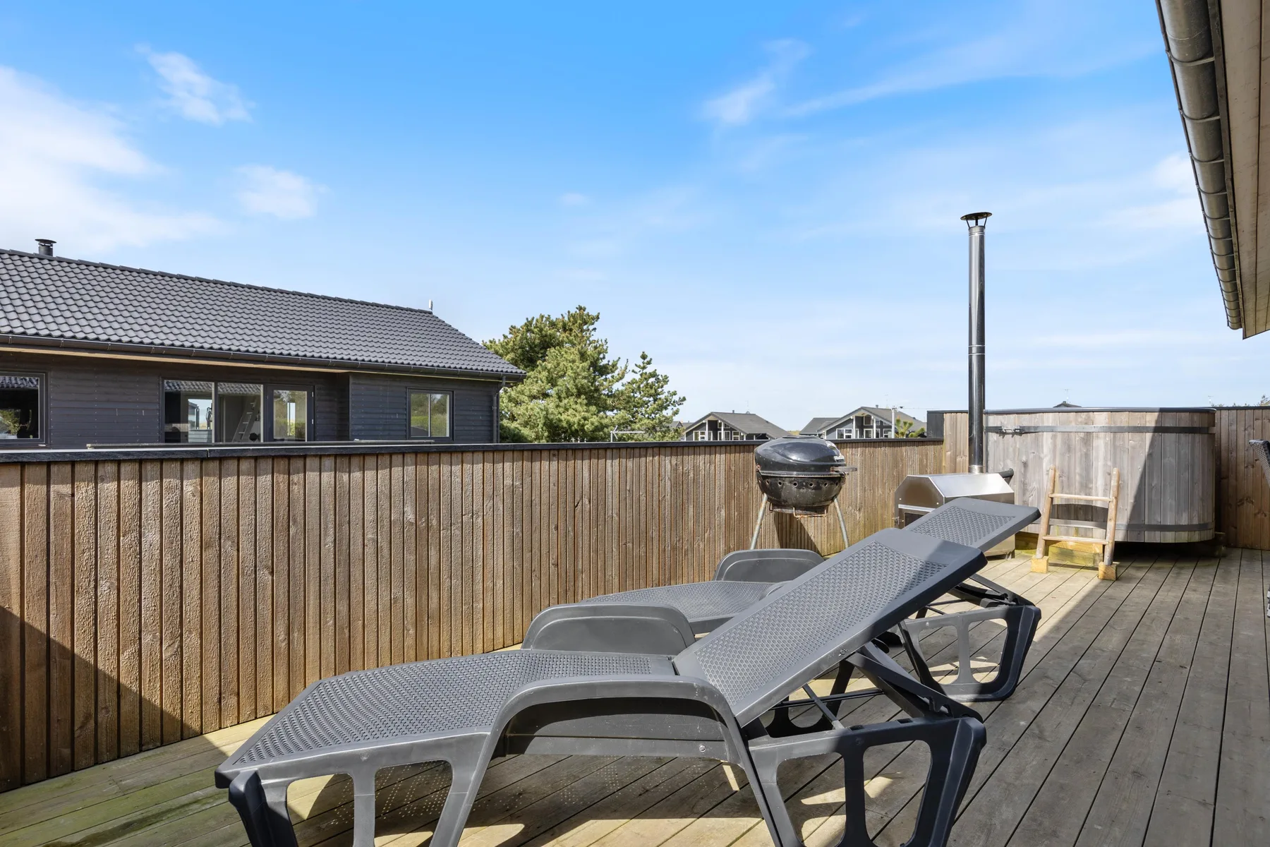 A wooden deck has reclining seating, a grill, and a fenced perimeter.