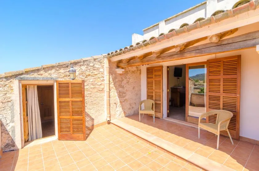 A sunlit patio with tiled flooring, wooden shutters, and seating outside a stone building.