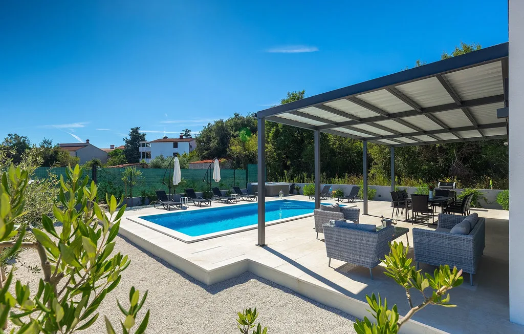 Una piscina al aire libre junto a un patio cubierto con asientos y muebles de comedor.