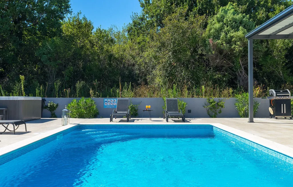 Una piscina al aire libre está rodeada por una terraza pavimentada y vegetación.
