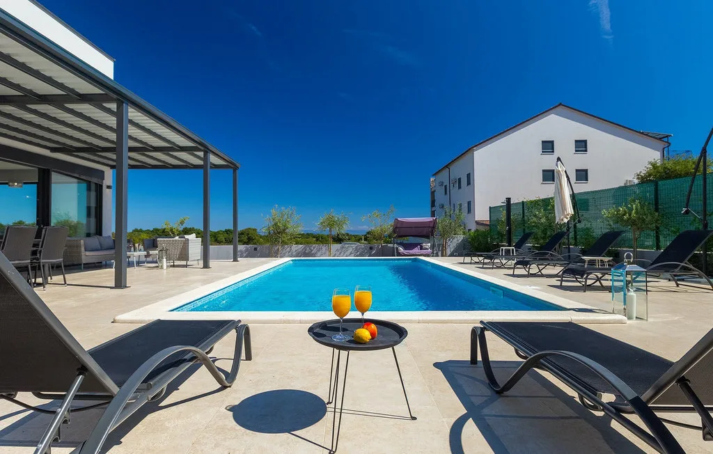 Una piscina al aire libre junto a una zona de estar con sombra, bajo un cielo despejado.
