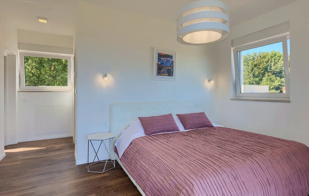 Un dormitorio con una cama doble cubierta con ropa de cama malva y ventanas con vistas a zonas verdes.