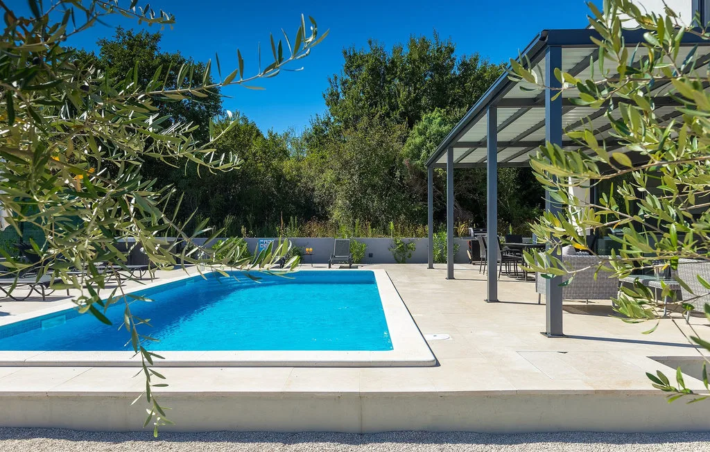 Una piscina al aire libre junto a una zona de asientos cubierta y árboles frondosos.