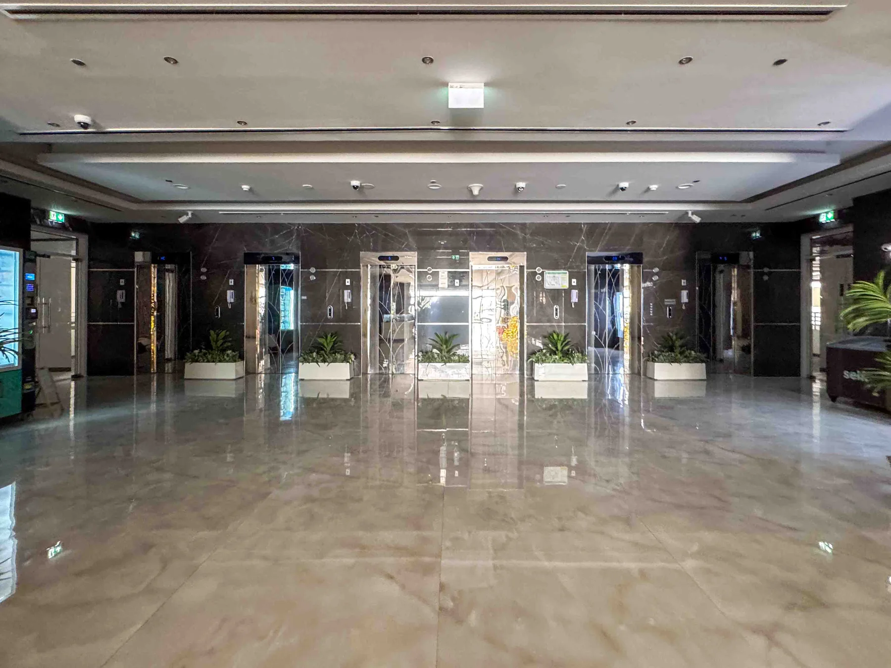 Un vestíbulo amplio con suelo reflectante, puertas de ascensor y jardineras junto a la pared del fondo.