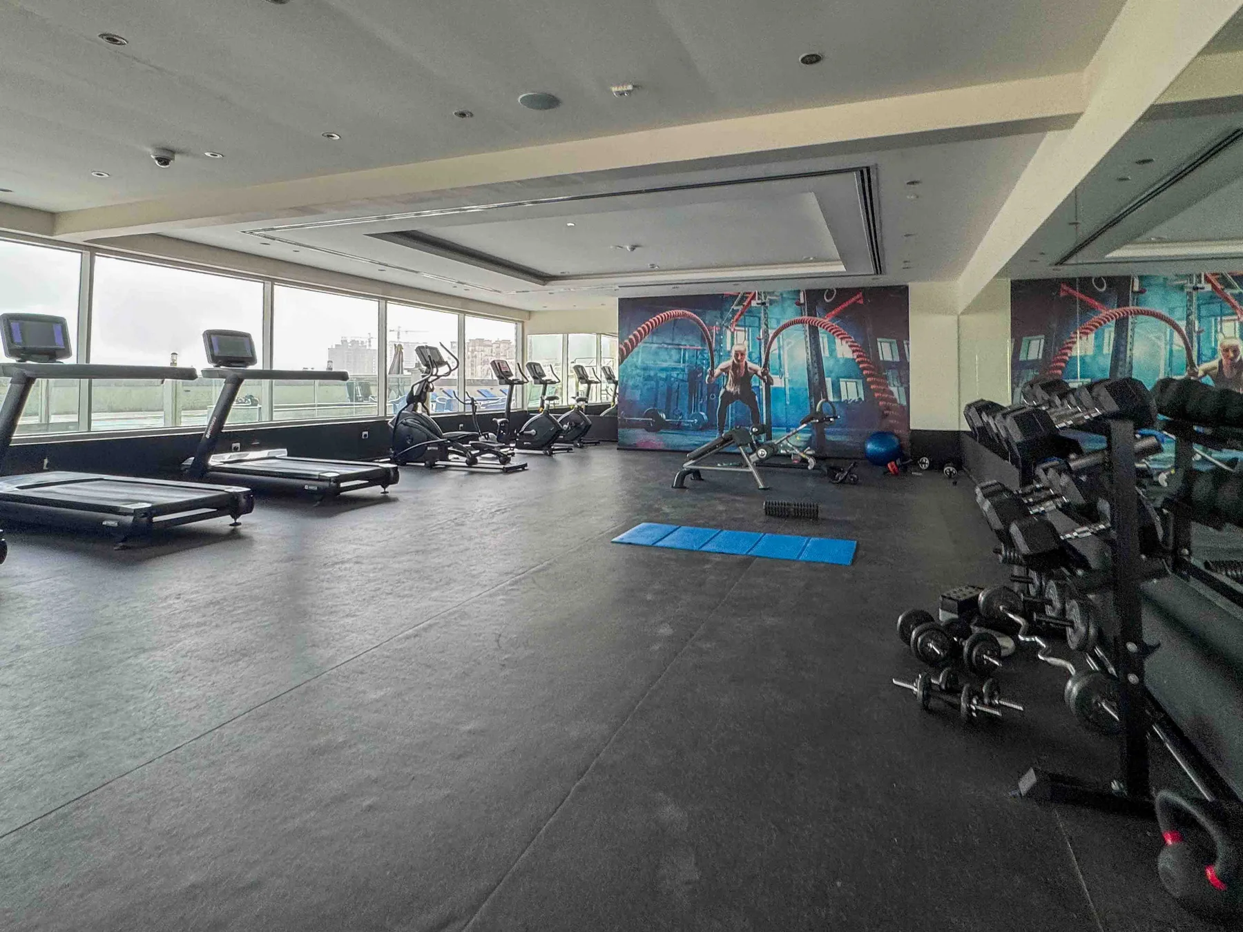 Un gimnasio con cintas de correr, bicicletas estáticas y pesas junto a grandes ventanas.