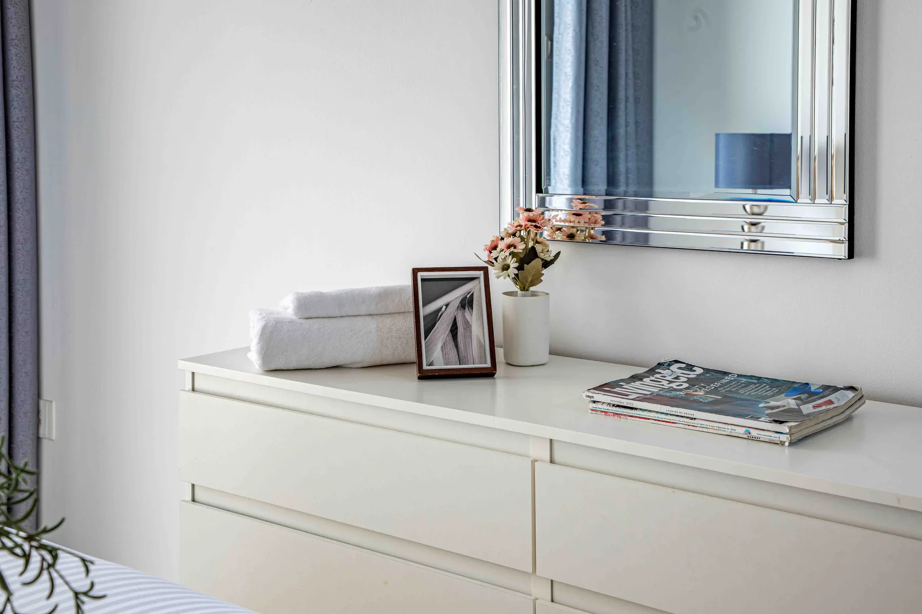 Una cómoda de dormitorio sostiene toallas dobladas, flores, una foto enmarcada y revistas bajo un espejo.