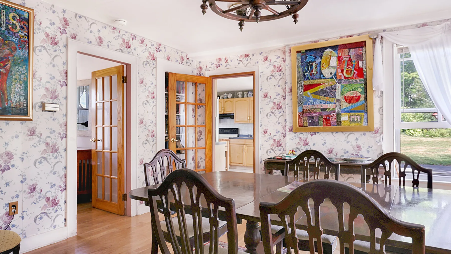 A dining room with dark wooden seating, floral wallpaper, and colorful framed artwork.