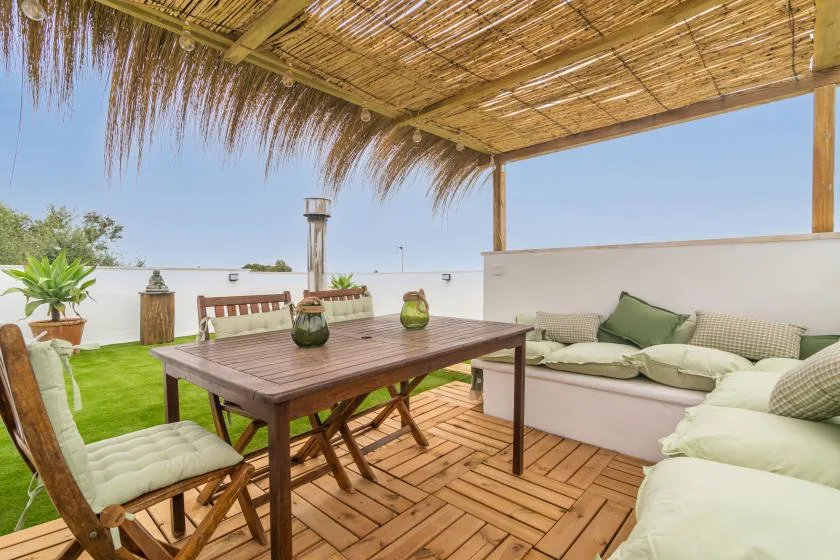 A shaded outdoor patio has a wooden dining table and cushioned seating.