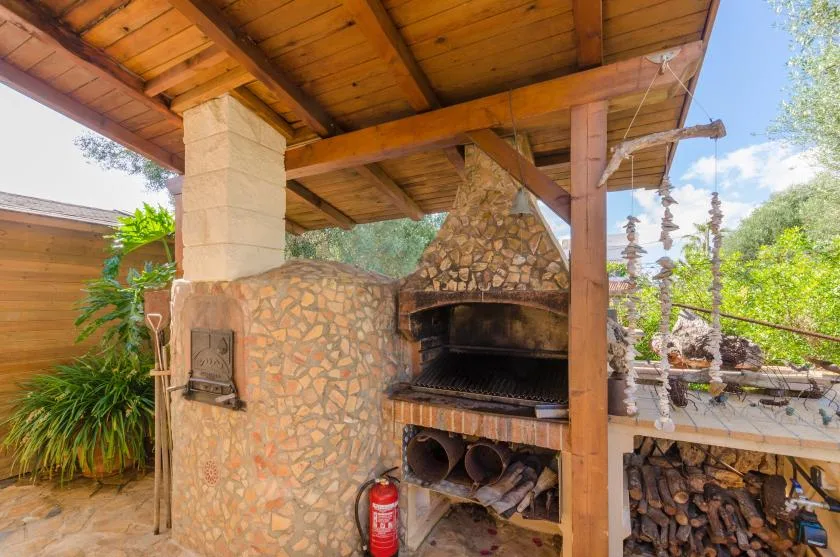 An outdoor stone barbecue beneath a wooden roof, with stacked firewood nearby.