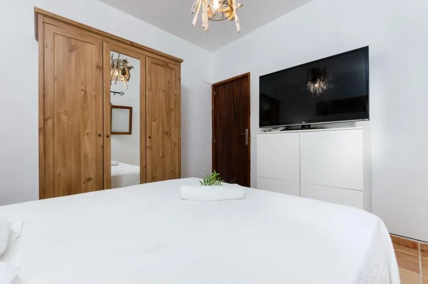 A bright bedroom with a white bed, wooden wardrobe, and wall-mounted television.