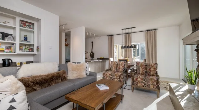 Una sala de estar con asientos, una mesa de centro de madera y grandes ventanas con cortinas.