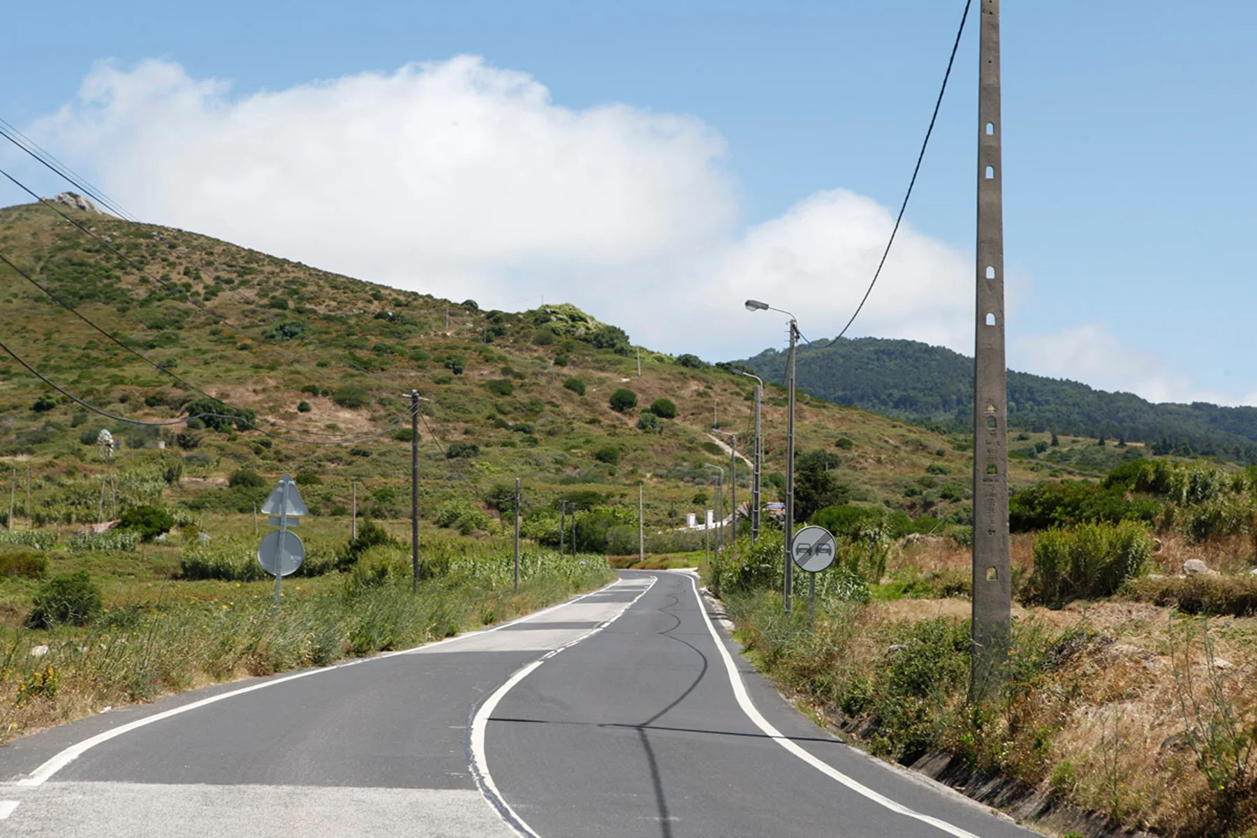 A paved road winds through dry, green hills beneath a partly cloudy sky.