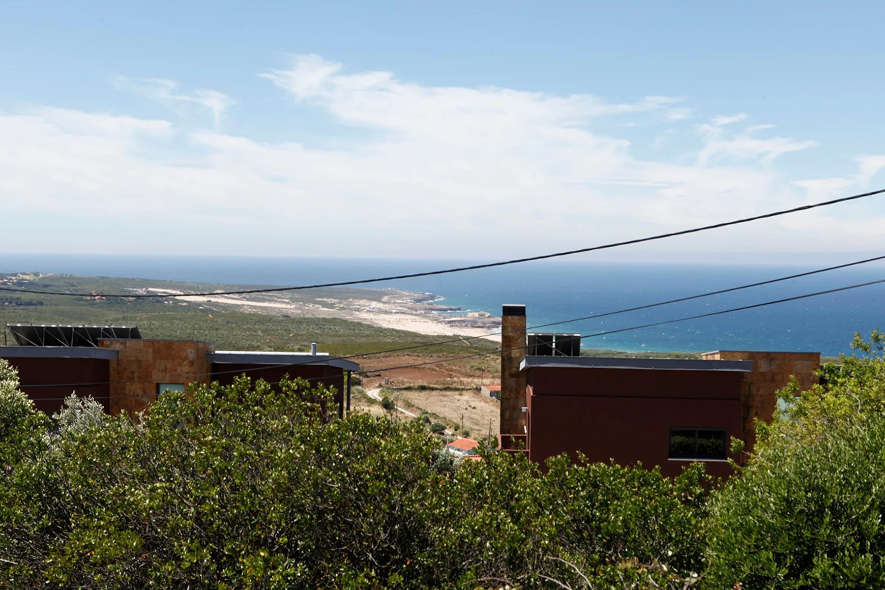 Coastal landscape with the ocean visible beyond low buildings and green shrubs.