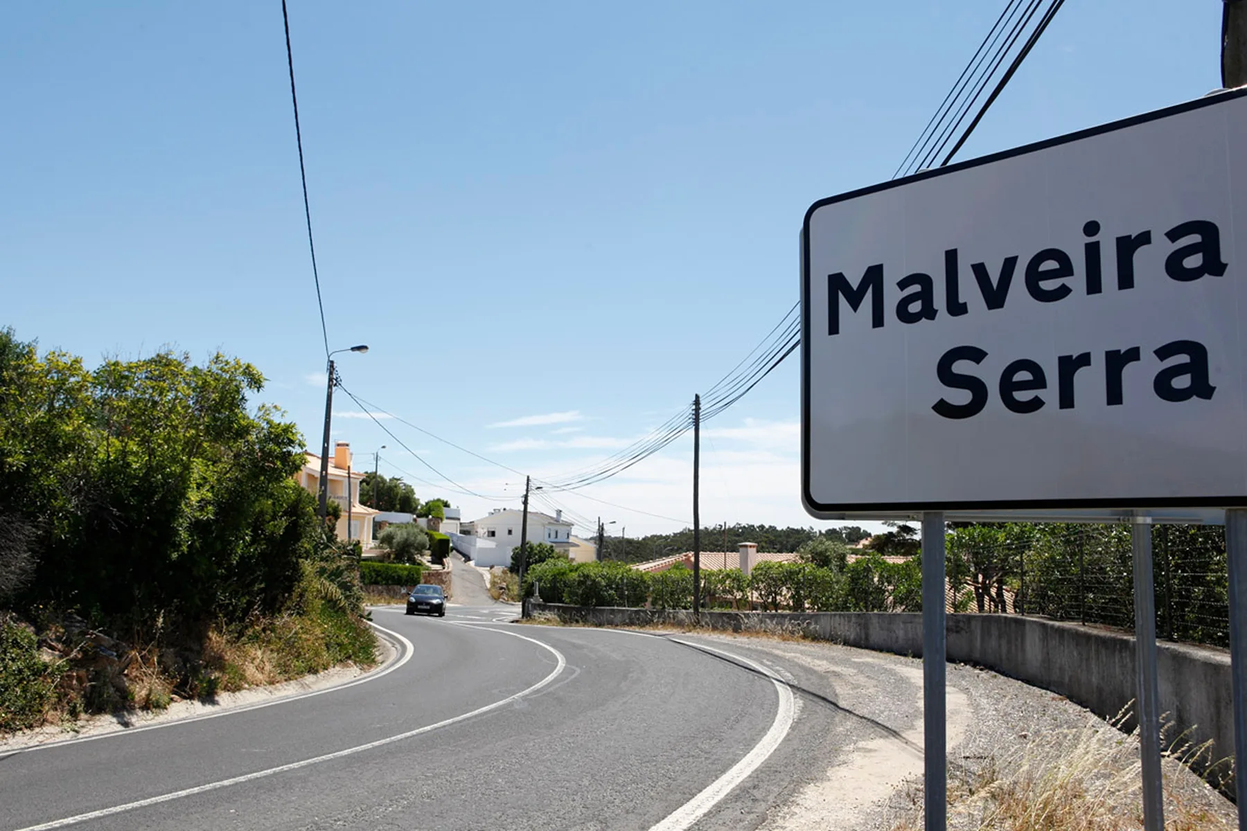 A paved road curves through a settlement beside a large Malveira Serra sign.