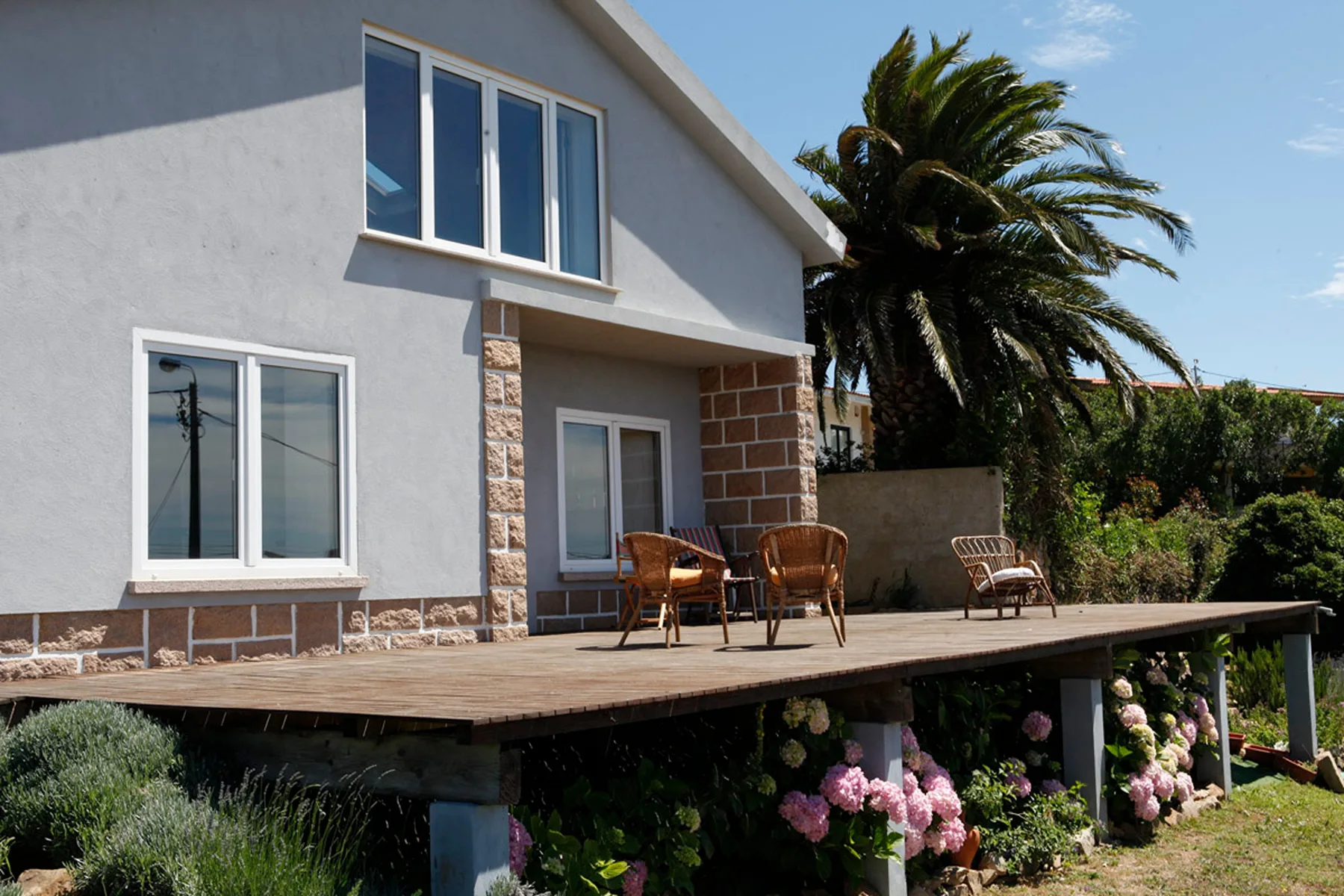 A house exterior opens onto a wooden deck with outdoor seating and flowering plants.
