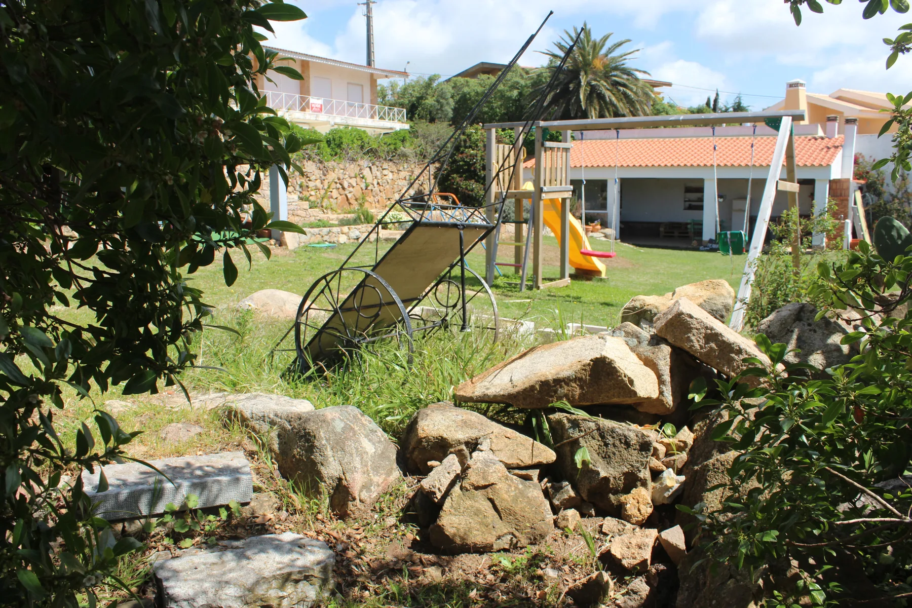 A garden surrounds a lawn with a swing, slide, and rocks in the foreground.