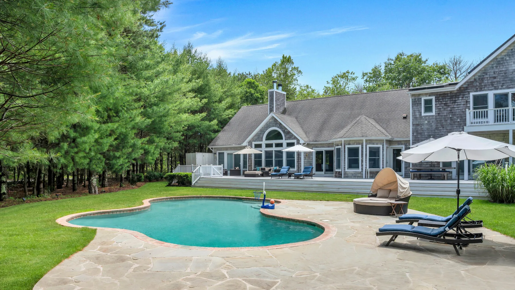 Una piscina exterior curva está junto a un amplio patio pavimentado y una casa grande.