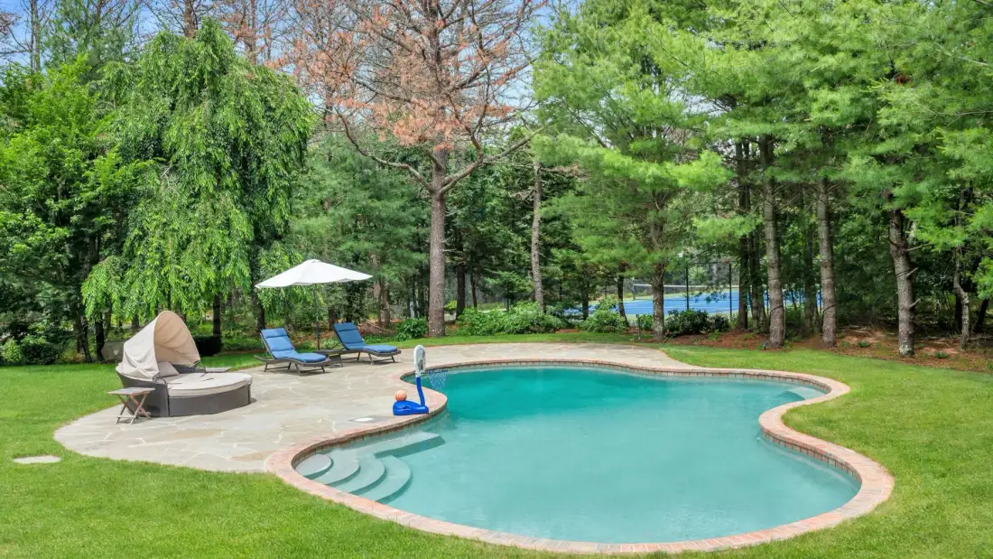 Una piscina curva está en un jardín con césped, rodeada de árboles.