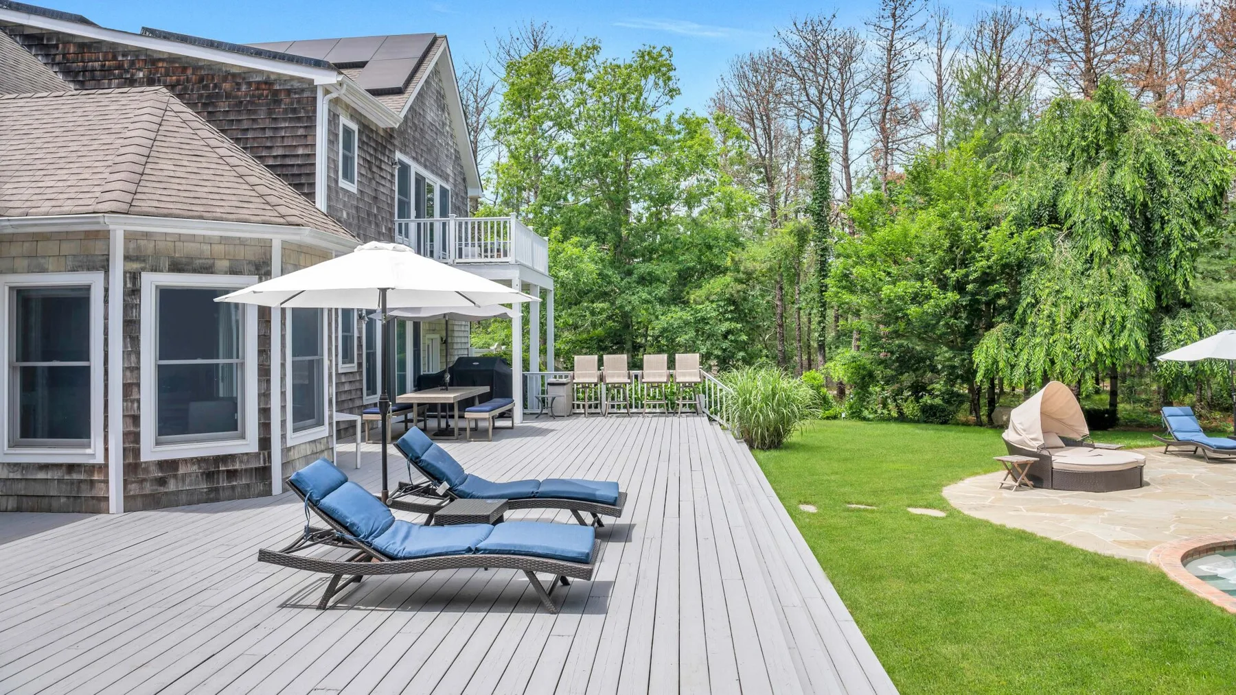 Una terraza exterior junto a una casa, con asientos reclinables y un patio con césped.